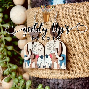 May include: A pair of wooden earrings with a dog mom design. The earrings are painted with brown, blue, white, and red paint. The earrings are shaped like a dog's head and have the words "Dog Mom" written on them.