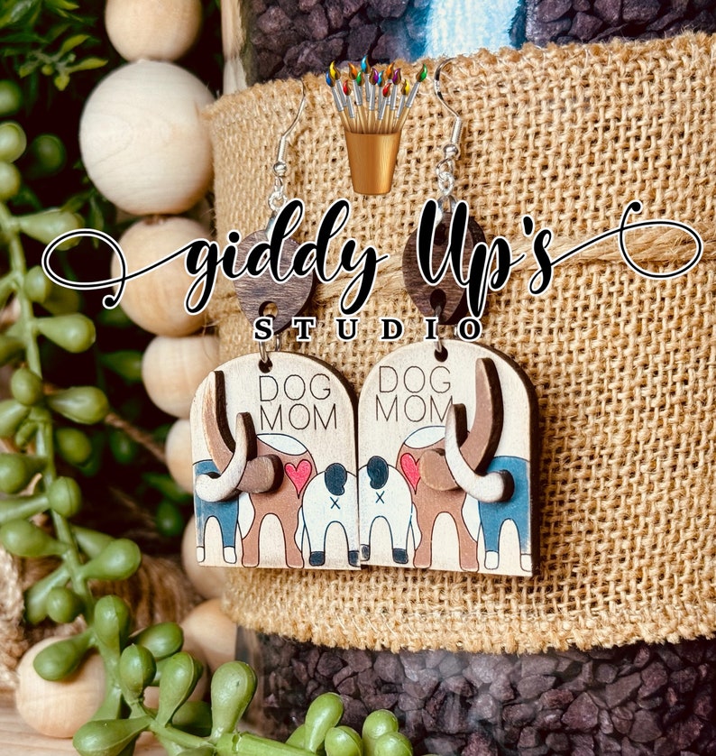 May include: A pair of wooden earrings with a dog mom design. The earrings are painted with brown, blue, white, and red paint. The earrings are shaped like a dog's head and have the words "Dog Mom" written on them.