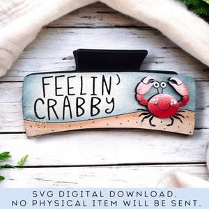 May include: A black hair clip with the words "FEELIN' CRABBY" and a cartoon crab illustration. The crab is red with pink claws and is on a beach scene with blue and tan colors.