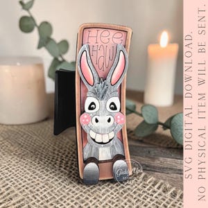 May include: A wooden donkey-themed SVG digital download design. The donkey has gray fur, pink ears and cheeks, and a large smile. The text "Hee Haw" is above the donkey. The design is on a wooden background.