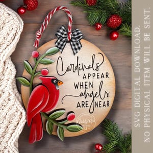 May include: Wooden round Christmas ornament with a red cardinal perched on a green branch with red berries. The ornament has the text "Cardinals Appear When Angels Are Near" and a black and white checkered bow.