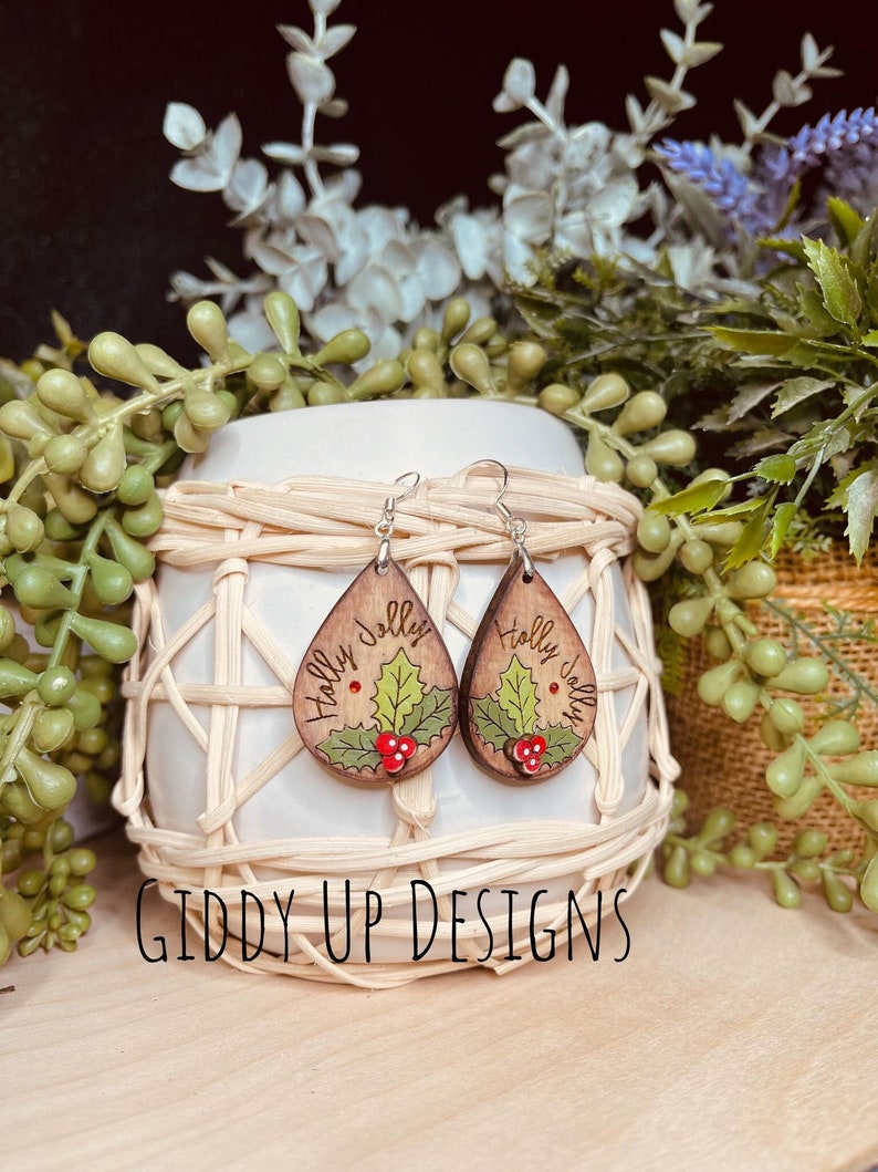 K&ouml;nnte beinhalten: Holz-Tropfenohrringe mit einem Stechpalmen-Design. Jeder Ohrring hat einen braunen Holzmaserungshintergrund mit einem gr&uuml;nen Stechpalmenblatt, roten Beeren und den Worten "Holly Jolly". Die Ohrringe werden auf einem geflochtenen Korb ausgestellt. Der Text "Giddy Up Designs" befindet sich unten.