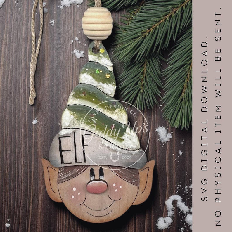May include: Wooden elf ornament with a green and white hat, brown hair, and a smiling face. The word "ELF" is printed on the hat. The ornament is hanging from a string with a wooden bead. The background is a wooden surface with a pine branch.