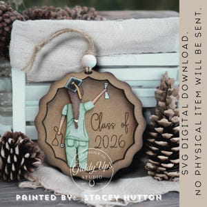 K&ouml;nnte beinhalten: Holzornament mit einer graduierenden Krankenschwester in OP-Kleidung, die einen Becher und ein Stethoskop h&auml;lt. Das Ornament tr&auml;gt die Aufschrift "Class of 2026" und stammt von Giddy Up's Studio. Der digitale SVG-Download wird gesendet, kein physischer Artikel.