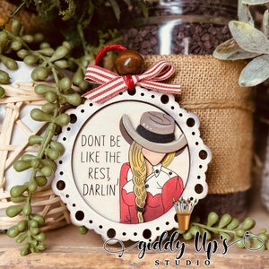 Puede incluir: Adorno de madera blanco con una cinta roja y blanca a rayas y una cuenta de madera marr&oacute;n. El adorno presenta una ilustraci&oacute;n de dibujos animados de una mujer con un sombrero de vaquero y una camisa roja con el texto "No seas como los dem&aacute;s, cari&ntilde;o" y las palabras "Giddy Up's Studio".