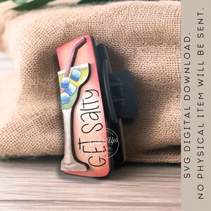 May include: A wooden hair clip featuring a cocktail design with the words "Get Salty." The clip has a black clasp and a colorful illustration of a cocktail with ice cubes and a straw. The background is a light pink.