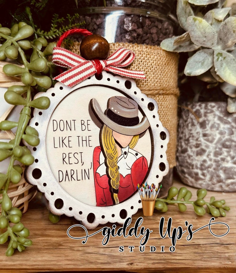 Puede incluir: Un adorno de madera blanco con un borde festoneado y una imagen pintada de una mujer con una camisa roja, un sombrero de vaquero y una trenza rubia. El texto "Don't be like the rest, darlin'" est&aacute; escrito en el adorno. El adorno est&aacute; decorado con una cinta roja y blanca y una cuenta de madera marr&oacute;n.