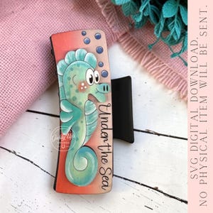 May include: A rectangular wooden sign featuring a cartoon seahorse in shades of teal and pink. The seahorse has a friendly expression and is surrounded by bubbles. The words "Under the Sea" are written vertically. The sign has a black stand.