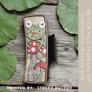 May include: A wooden frog and mushroom design. The frog is green with white eyes and sits on a red mushroom with white spots. The design is in a wooden frame. The text "Giddy Ups Studio" and "Painted by: Stacey Hutton" are visible.