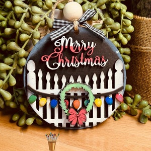 May include: A round wooden Christmas ornament with a black and white picket fence design. The ornament features the words "Merry Christmas" in pink and white lettering. A green wreath with a red bow is hanging on the fence, and colorful lights are strung across the top.
