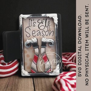 May include: A rectangular wooden sign with the text "It's Elf Season" and two elf legs. The sign is framed in black with a white border. The sign is decorated with red and white striped candy canes.