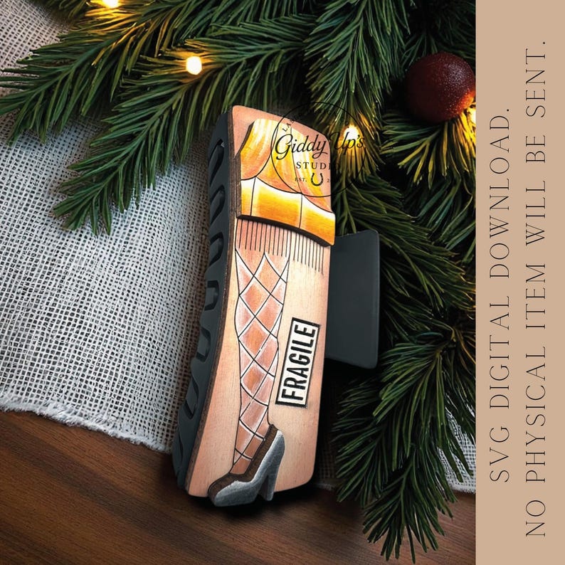 Puede incluir: Adorno navide&ntilde;o de madera con una ilustraci&oacute;n pintada de una pierna con medias de rejilla y tacones altos. El adorno tiene el texto "Giddy Up's Studio" y "FRAGILE". El fondo incluye ramas de pino.