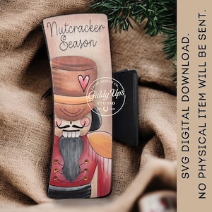 May include: Wooden sign featuring a painted nutcracker soldier with a brown hat, red jacket, and a large mustache. The sign reads "Nutcracker Season" and includes the text "Giddy Up's Studio". A black object is visible behind the sign.