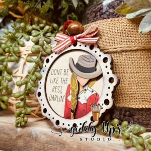 Puede incluir: Un adorno navide&ntilde;o de madera con una mujer que lleva una camisa roja y blanca y un sombrero de vaquero. El adorno tiene un fondo blanco con el texto "Don't be like the rest, darlin'" y las palabras "Giddy Up's Studio" en la parte inferior.