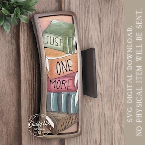 May include: A wooden hair clip featuring a stack of books with the words "JUST ONE MORE CHAPTER." The books are in various colors, including green, orange, and red. The clip has a dark brown clasp and a light brown frame.