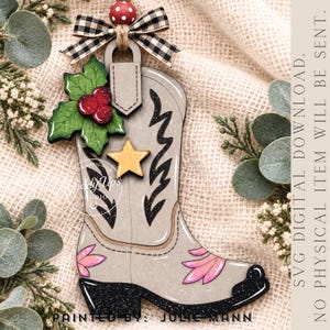 May include: A light gray cowboy boot ornament featuring a black glittery heel and toe, pink floral accents, a yellow star, and green holly leaves with red berries. A black and white checkered bow sits at the top with a red and white polka dot bead.