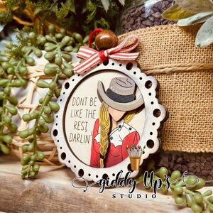 Puede incluir: Adorno de Navidad de madera con una mujer que lleva una camisa roja y blanca y un sombrero de vaquero. El adorno tiene un fondo blanco con el texto "Don't be like the rest, darlin'" y las palabras "Giddy Up's Studio" en la parte inferior.