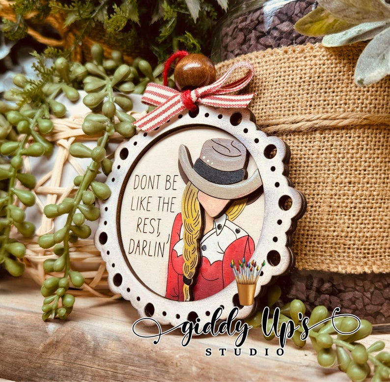 Puede incluir: Adorno de Navidad de madera con una mujer que lleva una camisa roja y blanca y un sombrero de vaquero. El adorno tiene un fondo blanco con el texto "Don't be like the rest, darlin'" y las palabras "Giddy Up's Studio" en la parte inferior.