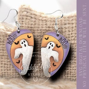 May include: A pair of Halloween-themed earrings. Each earring features a ghost design with the word "BOO!!!" and bats against a purple and orange background. The earrings are made by Giddy Up's Studio.