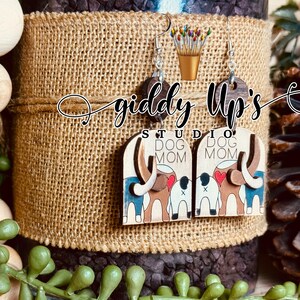 May include: A pair of wooden earrings with a dog mom design. The earrings are brown with a white background and feature a dog with a red heart and the words "Dog Mom".
