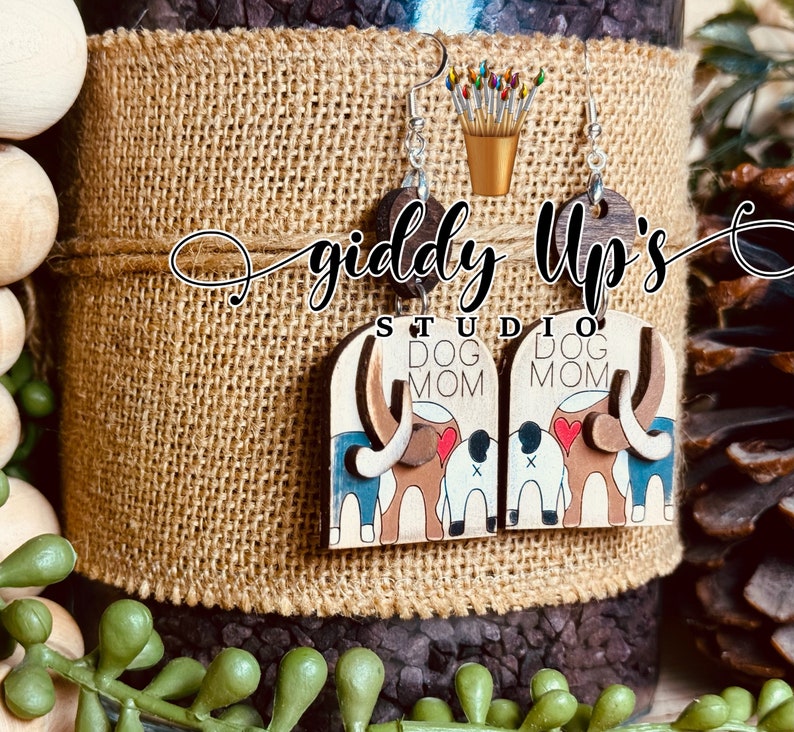 May include: A pair of wooden earrings with a dog mom design. The earrings are brown with a white background and feature a dog with a red heart and the words "Dog Mom".