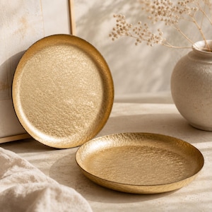 Könnte beinhalten: Zwei goldfarbene, runde Tabletts mit strukturierten Oberflächen. Die Tabletts haben ein gehämmertes Metallaussehen und einen dunklen Rand. Die Tabletts werden auf einer hellen Oberfläche mit einer Vase und Trockenblumen im Hintergrund präsentiert.