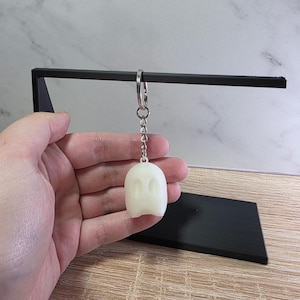 May include: A white ghost-shaped keyring with a silver chain. The keyring is held in front of a black display stand. The ghost has two oval eye cutouts. The background is a light grey and white marble pattern.