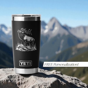 May include: Black stainless steel tumbler with a silver rim and a detailed etched image of a bull elk. The tumbler has the brand name "YETI" on the front. The background shows a mountain range.