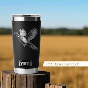 May include: Black stainless steel tumbler with a detailed engraving of a pheasant in flight. The tumbler has a silver rim and the brand name "YETI" is visible. The background is a blurred outdoor scene.
