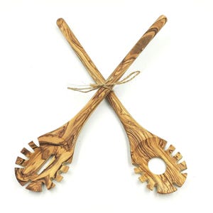 Op de afbeelding: Twee houten pastalepels van olijfhout, gekruist en vastgebonden met touw. De lepels hebben een natuurlijk nerfpatroon. Eén lepel heeft een gleufontwerp en de andere heeft een gat. Keukengerei.