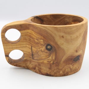 Può includere: Una tazza di legno con un design unico intagliato a mano. La tazza ha due fori nel manico per una presa facile. Il legno è di colore marrone chiaro con venature marroni più scure.