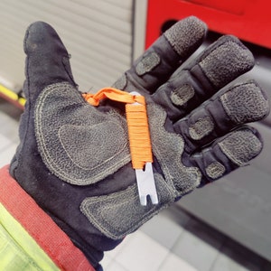 Peut inclure: Un gant de travail noir avec un outil orange et argenté attaché. L'outil a une petite boucle et un crochet en métal.