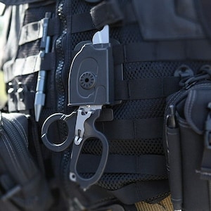 Op de afbeelding: Een zwart tactisch mes met een zwarte schede bevestigd aan een zwart tactisch vest. Het mes heeft een zilveren lemmet en een zwarte handgreep.