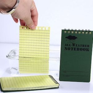 May include: A green all-weather notebook with a black cloud and raindrops graphic. The notebook has a black spiral binding and the text "ALL-WEATHER NOTEBOOK" printed on the front cover. The notebook is waterproof and tear-resistant.