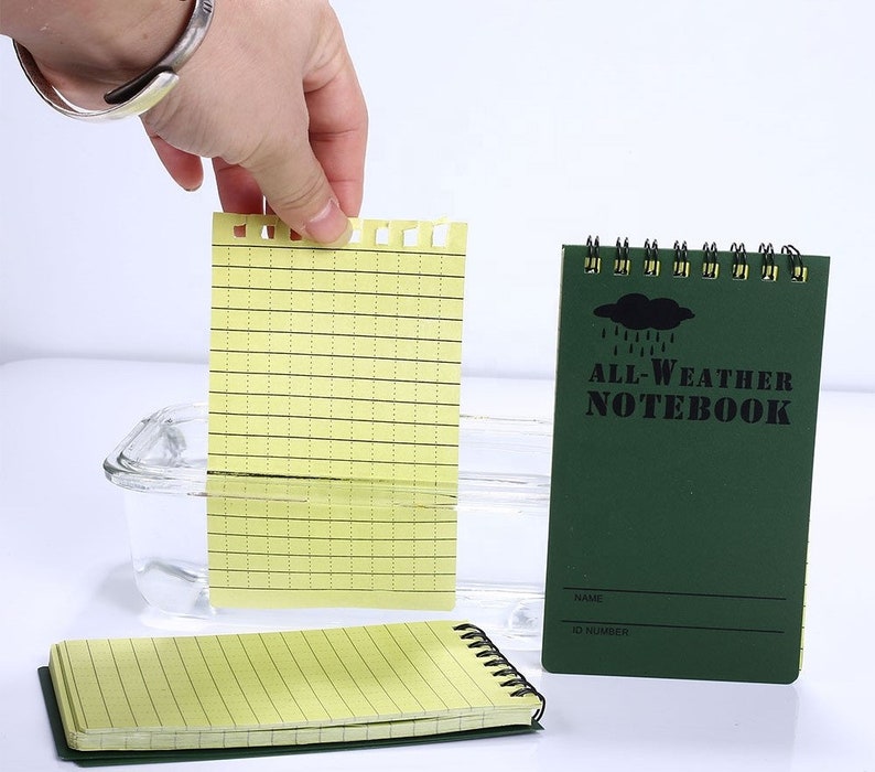 K&ouml;nnte beinhalten: Ein gr&uuml;nes Allwetter-Notizbuch mit einer schwarzen Wolken- und Regentropfengrafik. Das Notizbuch hat eine schwarze Spiralbindung und den Text "ALL-WEATHER NOTEBOOK" auf dem vorderen Umschlag. Das Notizbuch ist wasserdicht und rei&szlig;fest.