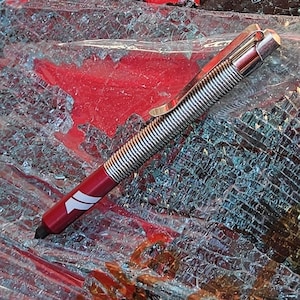 Op de afbeelding: Een zilveren en rode zelfverdedigingspen met een veermechanisme. De pen ligt op een gebroken stuk glas.