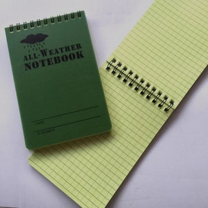 K&ouml;nnte beinhalten: Ein gr&uuml;nes Spiralnotizbuch mit dem Text "ALL-WEATHER NOTEBOOK" auf dem vorderen Umschlag. Das Notizbuch hat eine schwarze Wolke mit Regentropfen und Platz f&uuml;r einen Namen und eine ID-Nummer.
