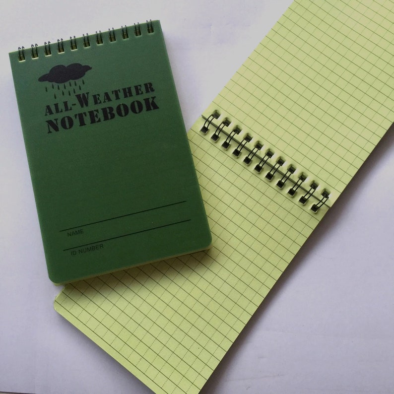 K&ouml;nnte beinhalten: Ein gr&uuml;nes Spiralnotizbuch mit dem Text "ALL-WEATHER NOTEBOOK" auf dem vorderen Umschlag. Das Notizbuch hat eine schwarze Wolke mit Regentropfen und Platz f&uuml;r einen Namen und eine ID-Nummer.
