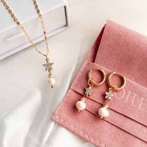 May include: A gold necklace and earrings set with a delicate flower charm and a pearl drop. The jewelry is displayed in a white box and a pink pouch.