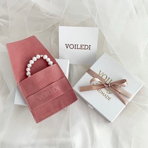 May include: A pink velvet jewelry pouch with a pearl bracelet inside. The pouch has the word "VOILEDI" embossed on it. The bracelet is in a white box with a pink ribbon and the words "VOILEDI Jewelry Handmade" on it.