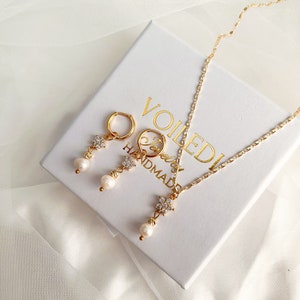 May include: A gold necklace and earrings set with a pearl drop and a flower design with clear rhinestones. The jewelry is displayed in a white box with the text "Voiledi Handmade".