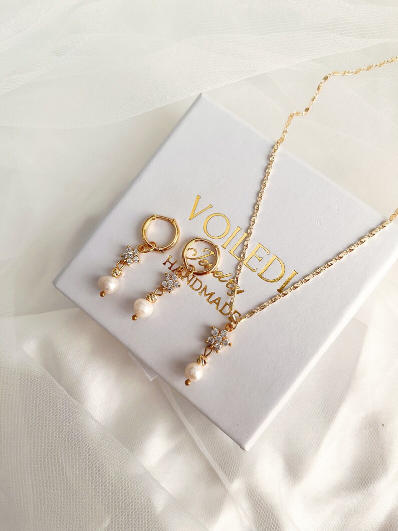 May include: A gold necklace and earrings set with a pearl drop and a flower design with clear rhinestones. The jewelry is displayed in a white box with the text "Voiledi Handmade".