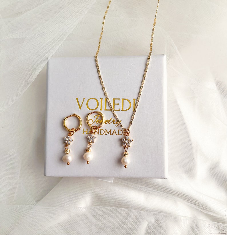 May include: A gold necklace and earrings set with a delicate flower charm and a single pearl drop. The jewelry is displayed on a white box with the text "VOILEDI Jewelry HANDMADE".
