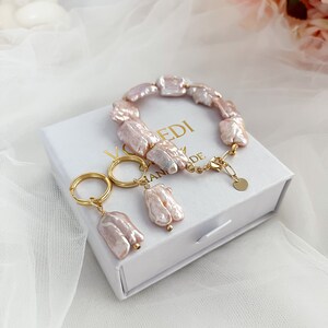 Pu&ograve; includere: Un set di gioielli delicati composto da un braccialetto e orecchini realizzati con perle d'acqua dolce rosa e dettagli dorati. I gioielli sono esposti in una scatola bianca con la scritta "HANDMADE" su di essa.