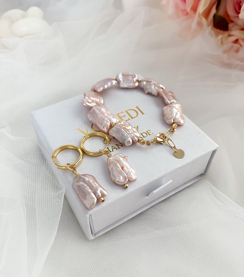 Pu&ograve; includere: Un set di gioielli delicati composto da un braccialetto e orecchini realizzati con perle d'acqua dolce rosa e dettagli dorati. I gioielli sono esposti in una scatola bianca con la scritta "HANDMADE" su di essa.