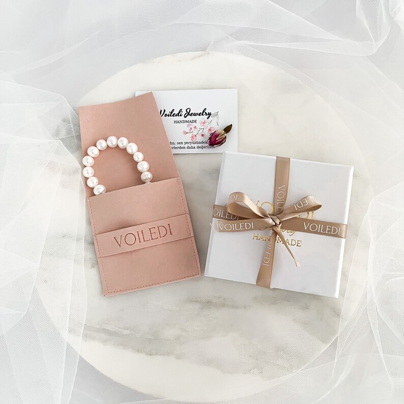 Pu&ograve; includere: Un braccialetto di perle bianche in una pochette di velluto rosa con la scritta "VOILEDI" stampata su di essa. La pochette &egrave; appoggiata su una superficie di marmo bianco con una scatola regalo bianca con un nastro dorato e la scritta "VOILEDI" stampata su di essa.