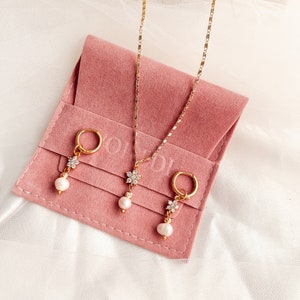 May include: A set of gold jewelry featuring a delicate flower pendant necklace and matching earrings with pearl drops. The jewelry is displayed on a pink velvet pouch.