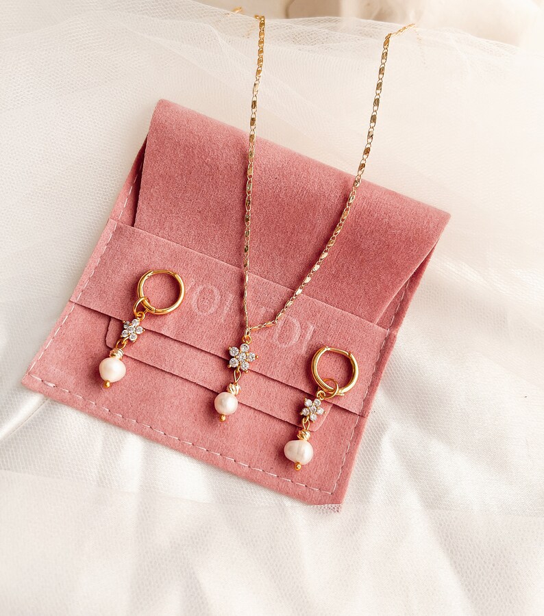 May include: A set of gold jewelry featuring a delicate flower pendant necklace and matching earrings with pearl drops. The jewelry is displayed on a pink velvet pouch.