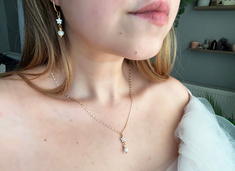 May include: A gold chain necklace with a small pearl and flower pendant. The necklace is paired with matching earrings with a pearl and flower design.