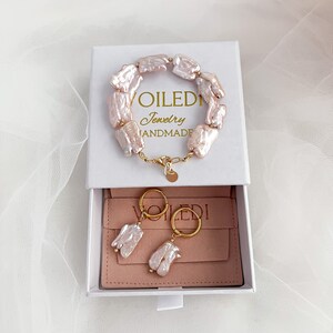 Pu&ograve; includere: Una scatola bianca con un interno rosa che contiene un braccialetto e orecchini di perle. Il braccialetto e gli orecchini sono in oro e hanno un design a forma di perla. La scatola ha il testo "VOILEDI Jewelry HANDMADE" sul coperchio.